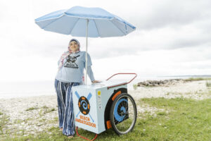 Woman standing under an umbrella that is attached to a wheeled cart near water. 