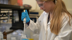 Blonde woman wearing safety goggles, gloves and a lab coat in a lab. 
