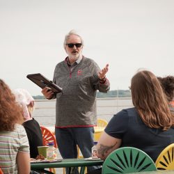Standing man wearing sunglasses talking with seating listeners and a lake in the background.