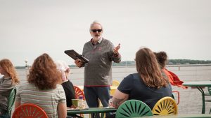 Standing man wearing sunglasses talking with seating listeners and a lake in the background. 