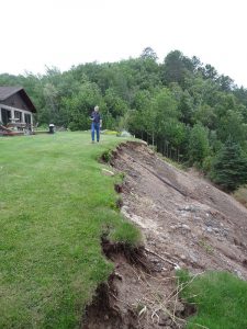 Person walking along a tall bluff that is highly eroded, with a house perched dangerously above the erosion