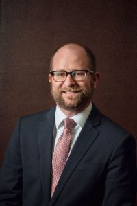 Close-up of a person wearing glasses, suit, tie and smiling. 