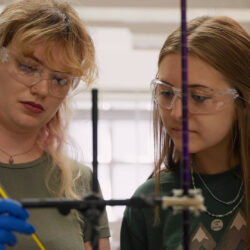 Close-up of two people in a lab, wearing safety goggles and one with blue lab gloves