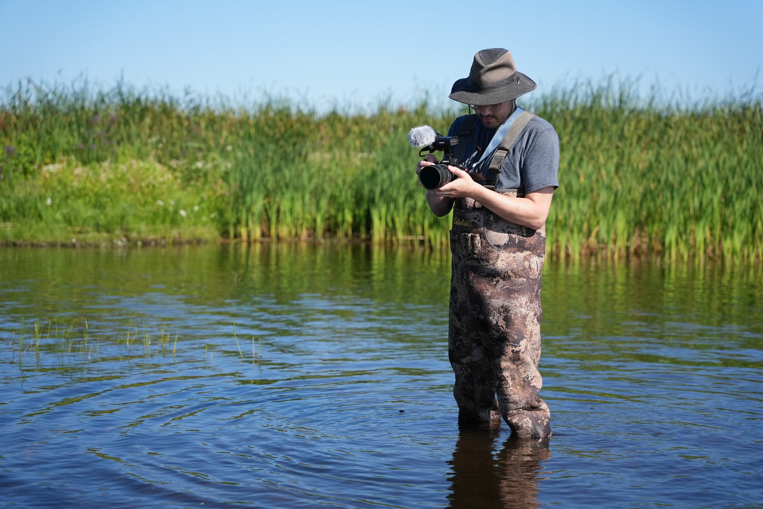Jeremy Van Mill stands in waders in a wetland, holding a camera.