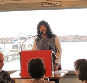 Person standing at a podium with water and a boat in the background.
