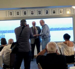 In the background, two people holding an award. A photographer standing in the foreground. 