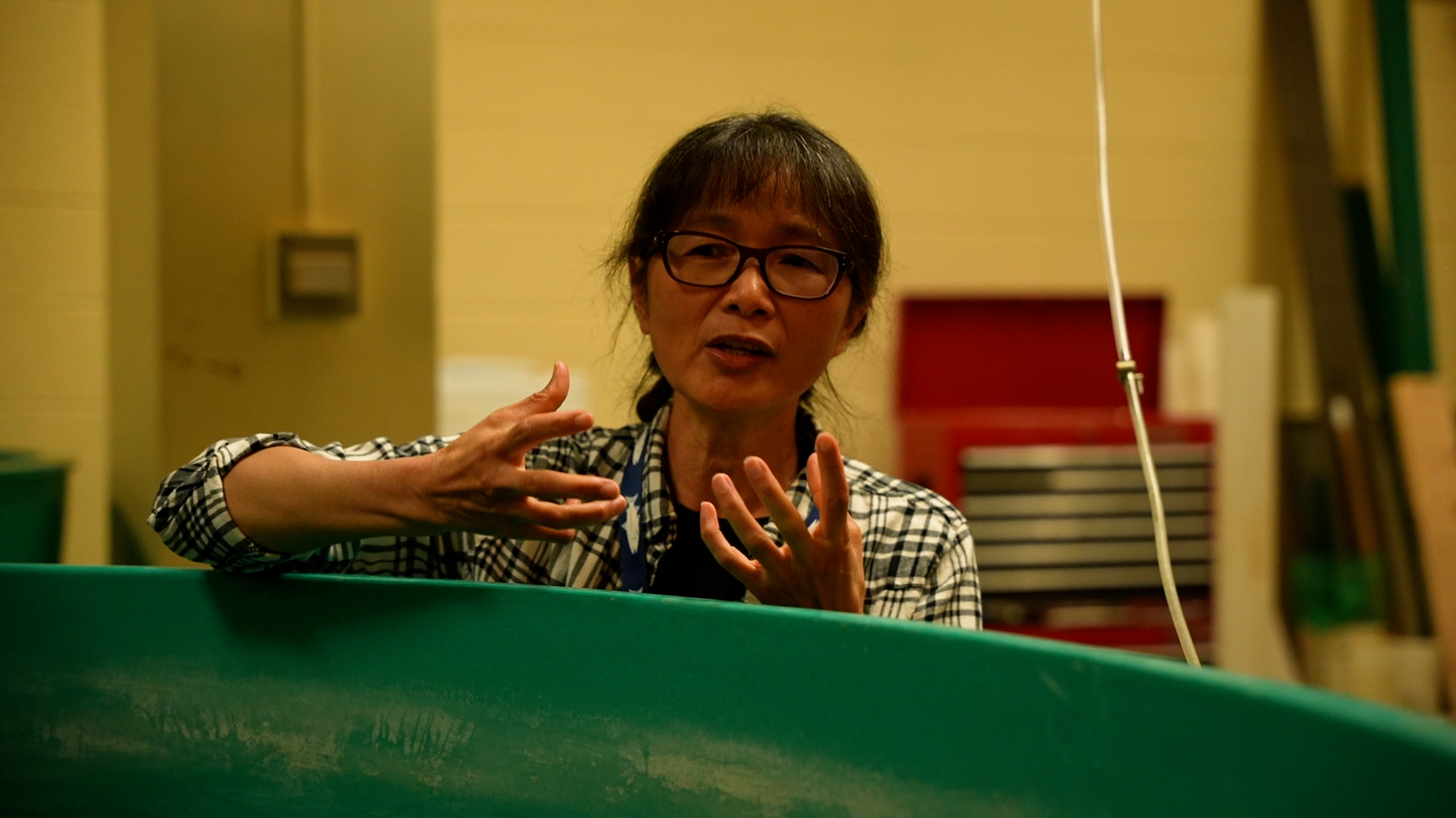 Aquaculture Outreach and Extension Specialist Dong-Fang Deng discusses stands behind a teal-colored fish tank in her lab.