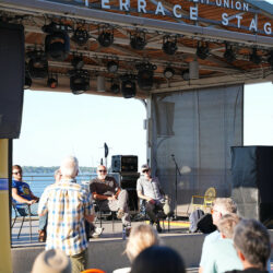 Stage with water in background. People sitting near stage and four people on the stage.