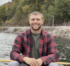 Person wearing a plaid shirt and smiling with water in the background. 