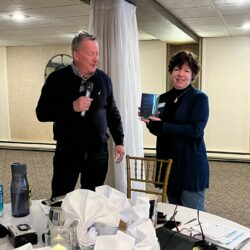 Two people standing next to each other in a room. One person is holding a microphone. The other person is holding an award.