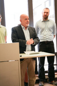 Person wearing a suit jacket standing at a podium with clasped hands. A second tall person stands in the background. 