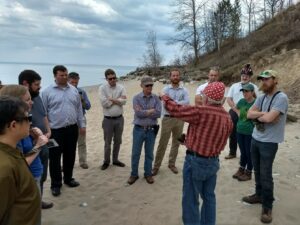 Large group of people standing on a beach with a lake in the background and a high bluff to the right. 