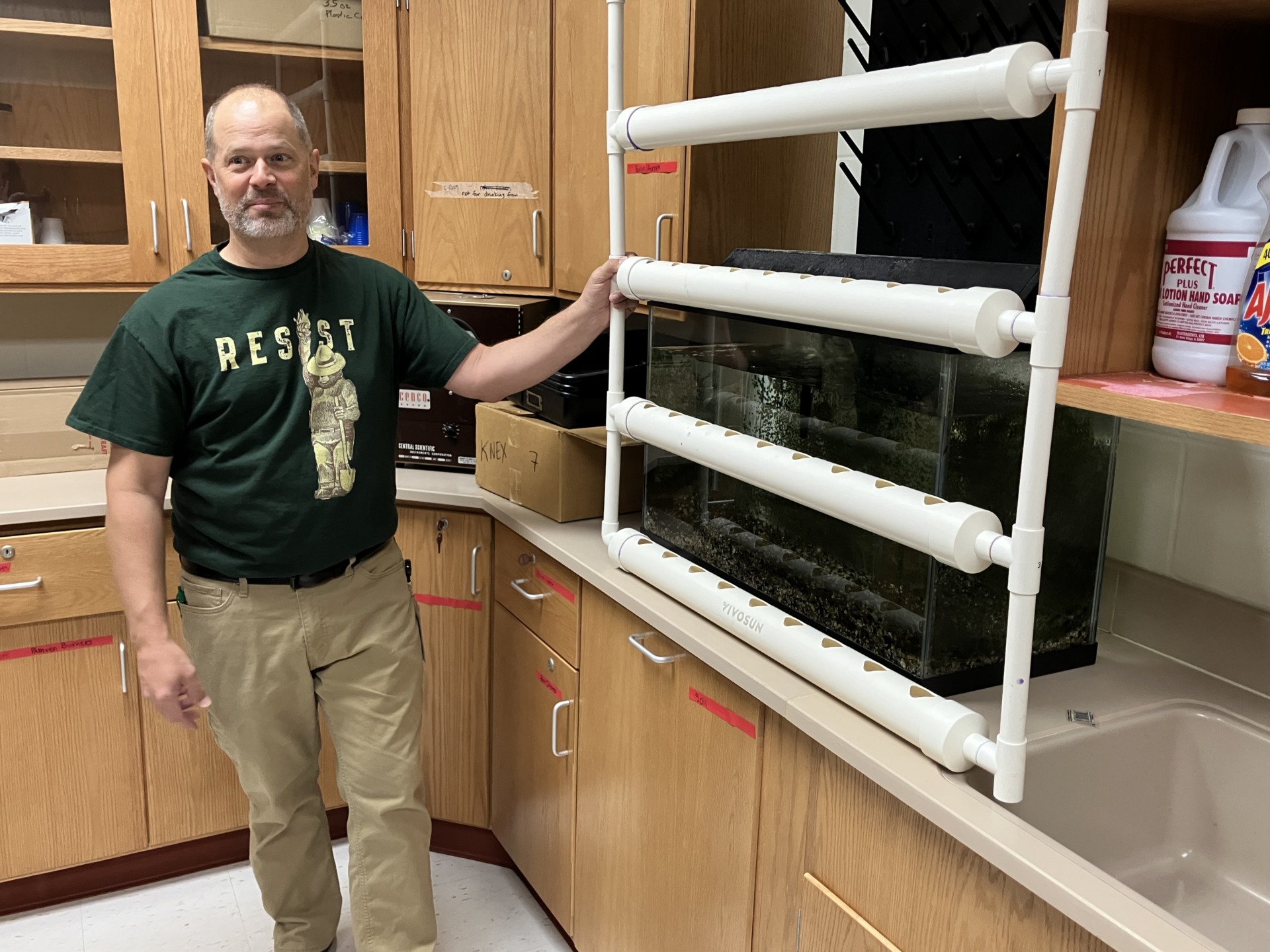 8th grade science teacher Dan Widiker stands next to a PVC-pipe rack for growing plants in an aquaponic system