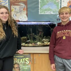 Two middle school students, Ava and Liam, pose for a photo on either side of the guppy tank in Mr. Widiker's 8th grade science class.