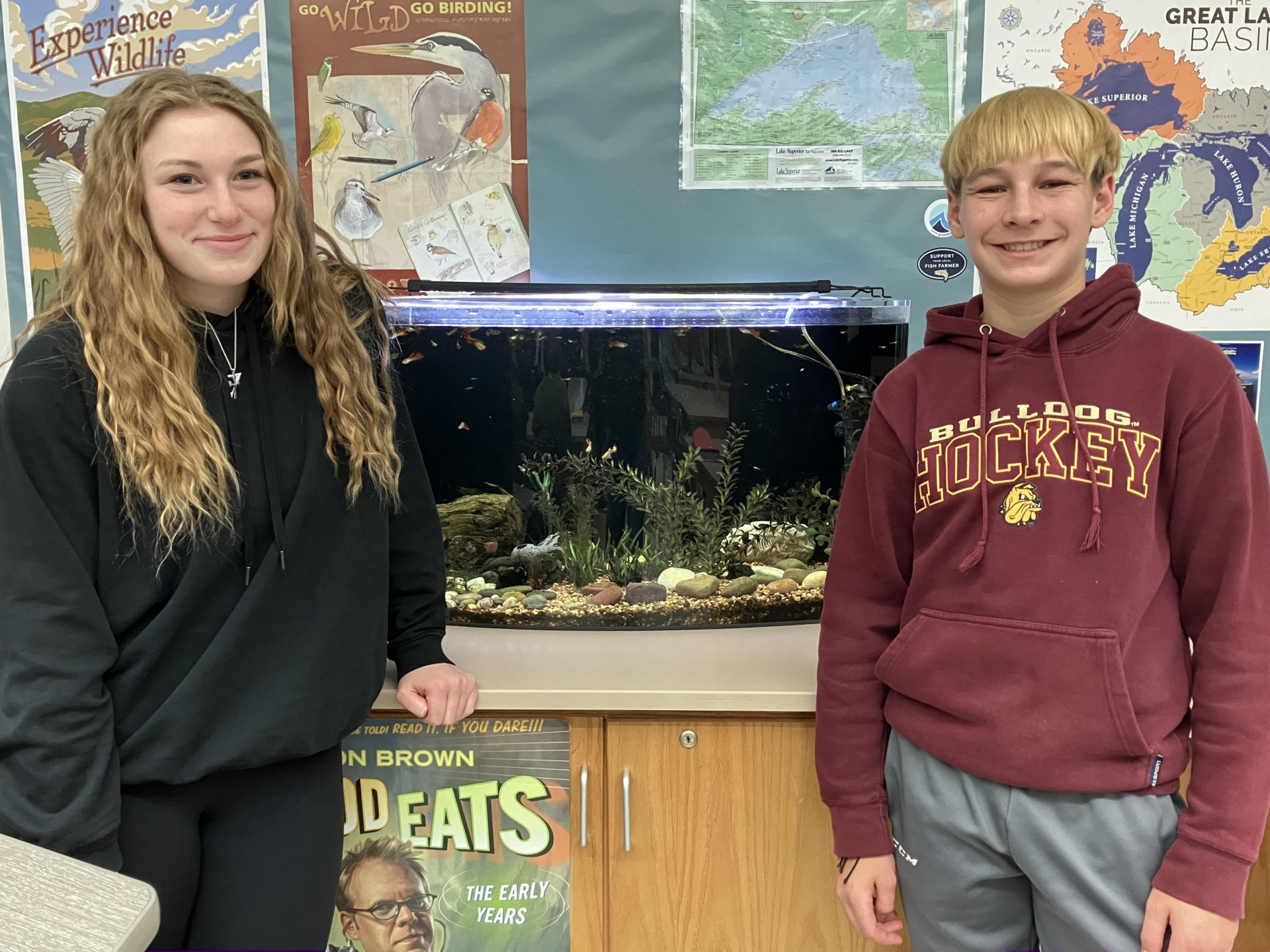 Two middle school students, Ava and Liam, pose for a photo on either side of the guppy tank in Mr. Widiker's 8th grade science class.