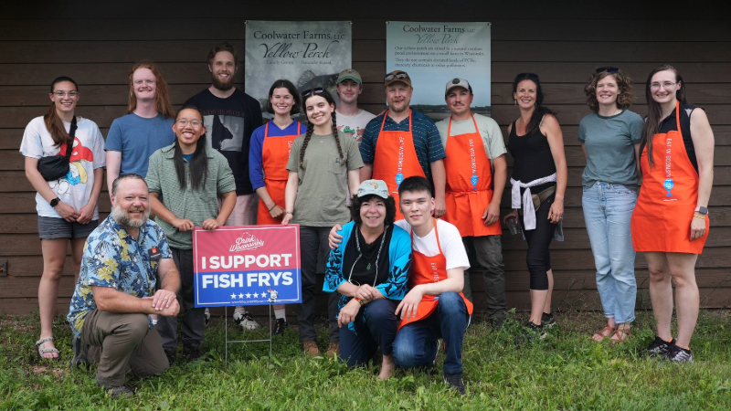 Group of interns and staff smile and gather around a sign saying "I support fish frys.'"
