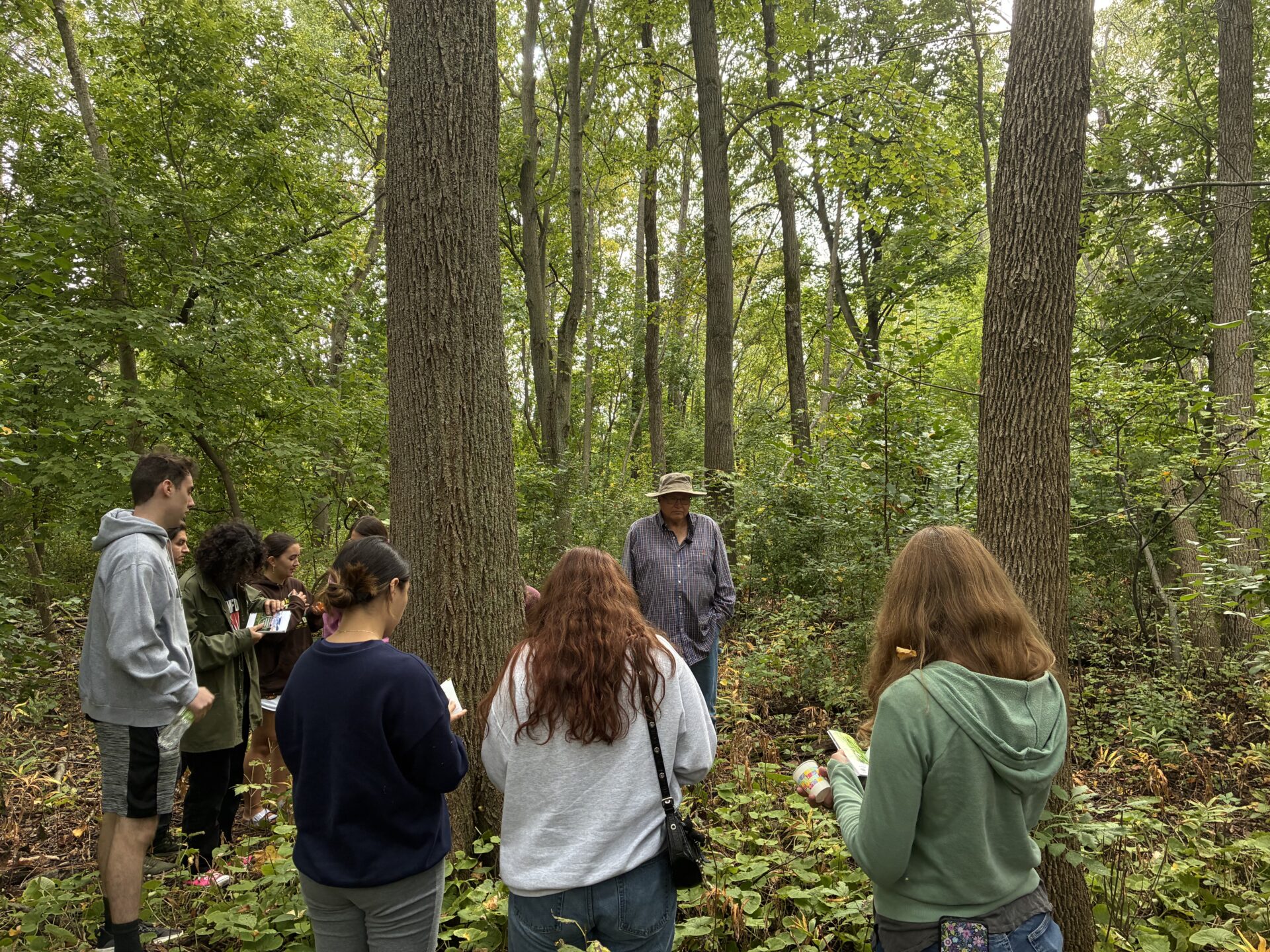 A group of UW–Green Bay students and staff gather around a man in a blue shirt in the woods.