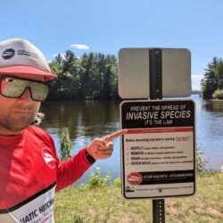 A man in reflective sunglasses wearing a Stop Aquatic Hitchikers shirt points to a sign telling boaters to remove invasive species from their boats