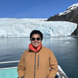 Marian Azeem-Angel wears a brown jacket and stands on a boat overlooking a glacier in Alaska