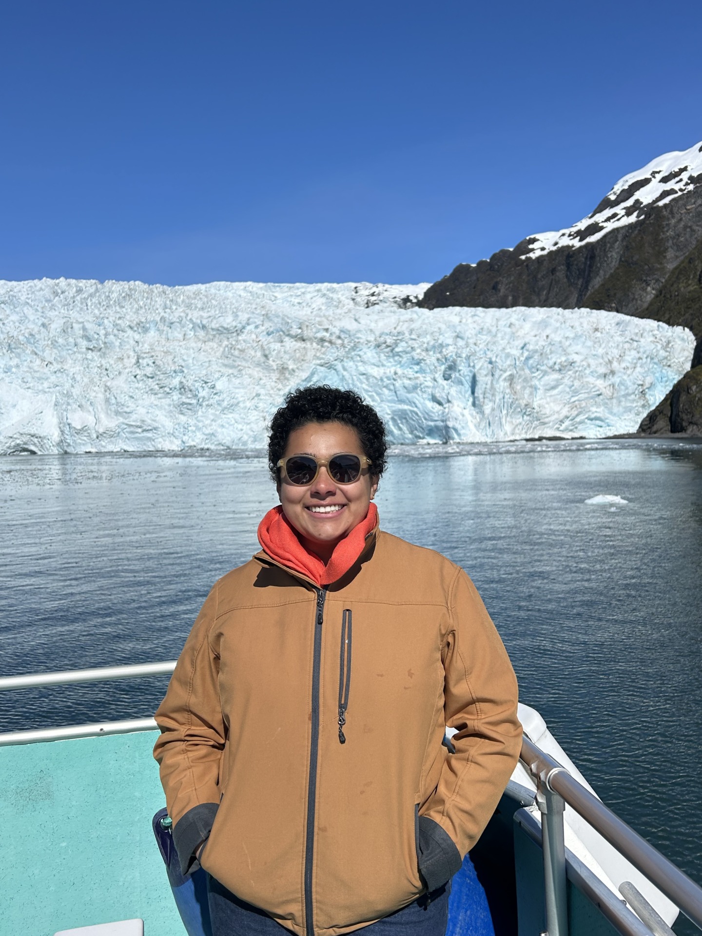 Marian Azeem-Angel wears a brown jacket and stands on a boat overlooking a glacier in Alaska