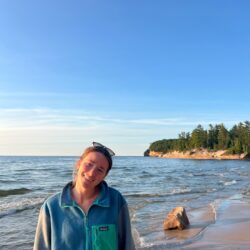 Cailin Young wears a blue jacket and stands on a Lake Superior beach