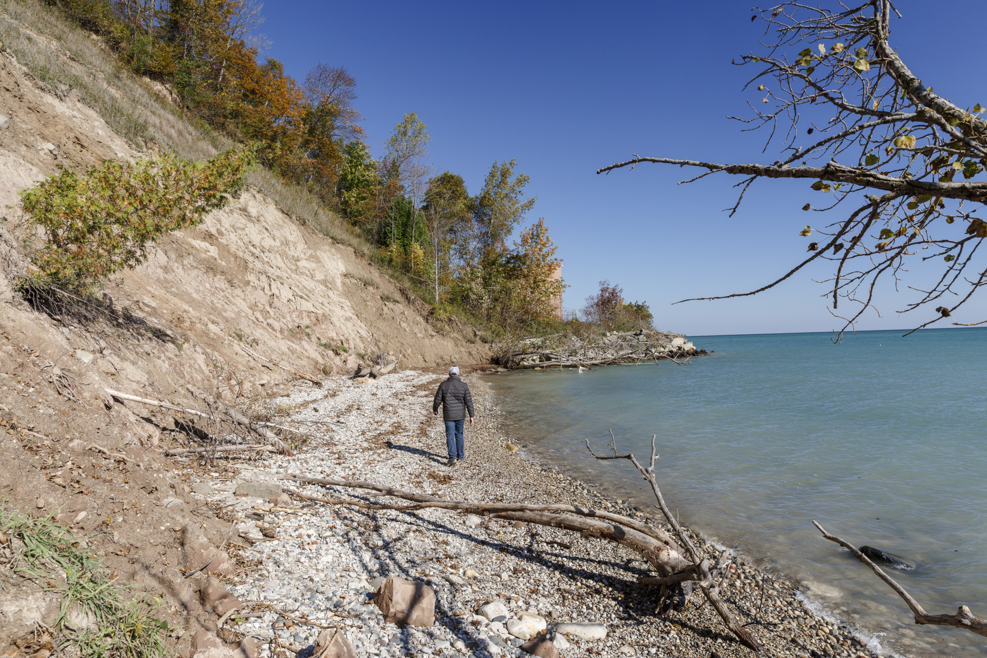 A person in a black jacket walks along a sandy, eroding bluff on Lake Michigan
