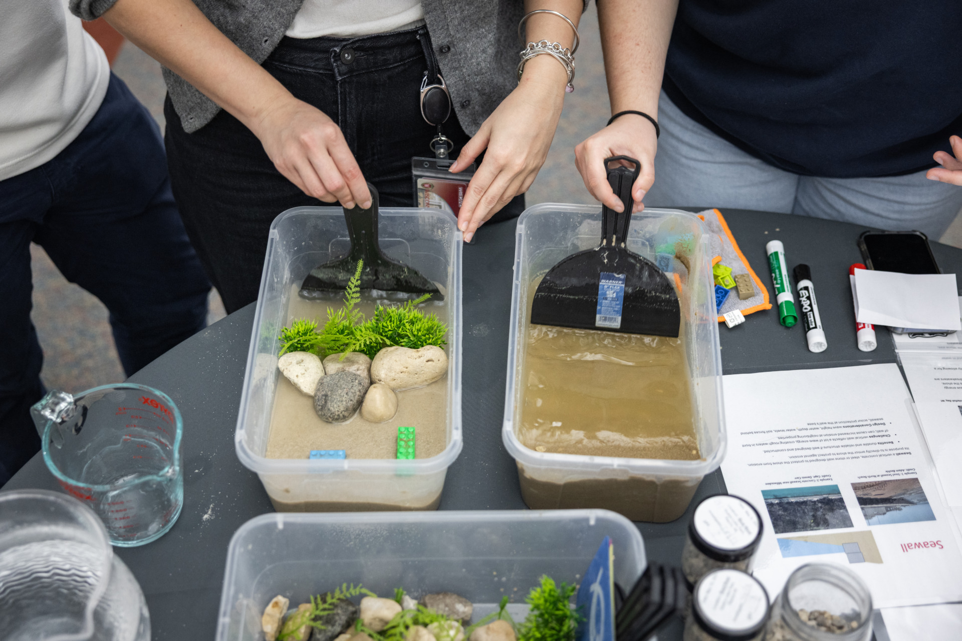 Students experiment with two wave tanks, plastic containers, about the size of a shoe box with sand and water. One contains rocks and plastic vegetation.