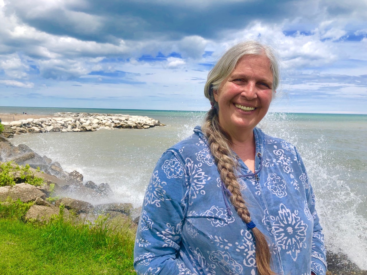 A woman with grey hair in a long braid wearing a blue shirt stands in front of Lake Michigan