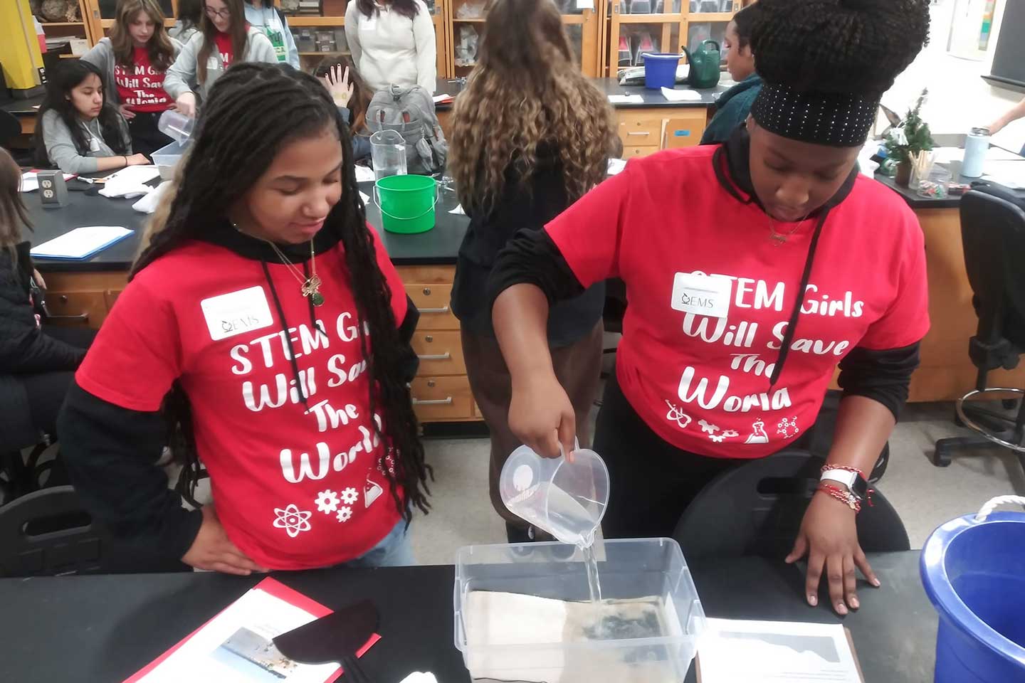 Two students experiment with a wave tank. The tank is a shoebox-sized plastic container filled with sand and water.